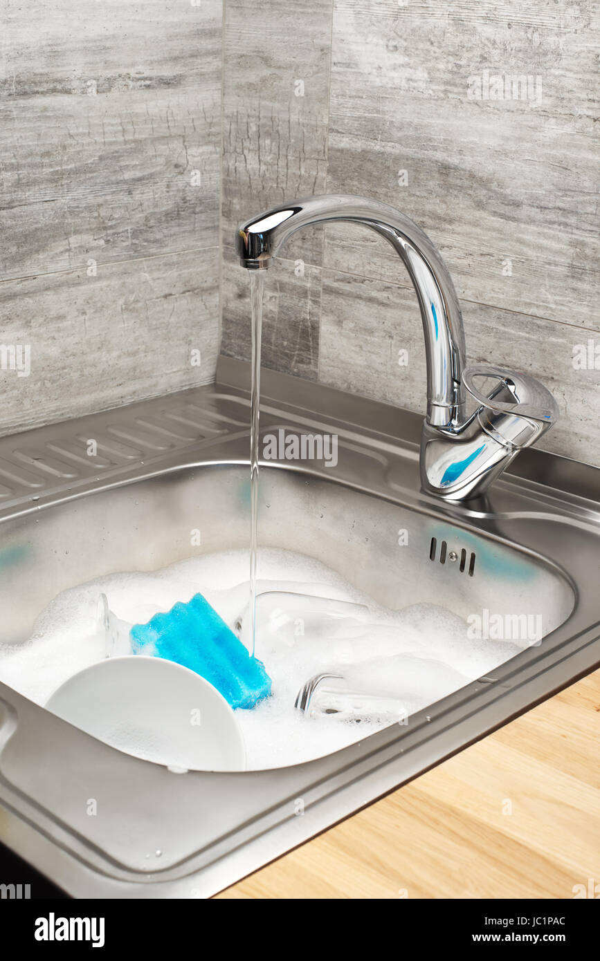 Nahaufnahme von Spülbecken mit fließendem Leitungswasser voller Schaum, Geschirr und Reinigungsschwamm blau Stockfoto