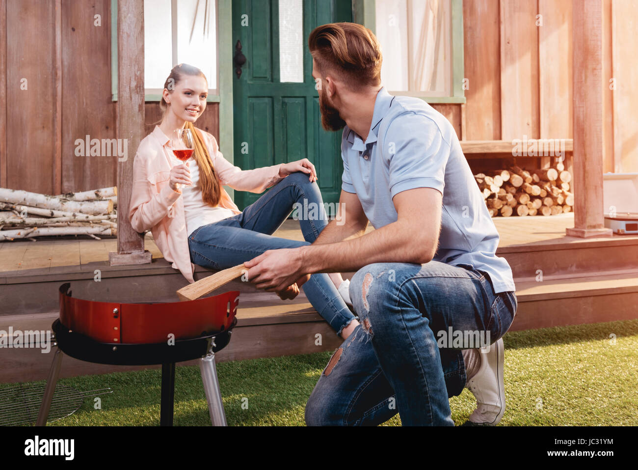 Lächelnde junge Frau trinken Wein und Blick auf Mann kniet in der Nähe von Grill im freien Stockfoto