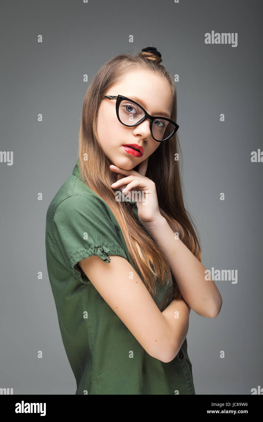 junge hübsche Brünette brillentragende Mädchen auf grauem Hintergrund. Frau in grün T-shirt und schwarze Jeans gekleidet. rote Lippen. Stockfoto