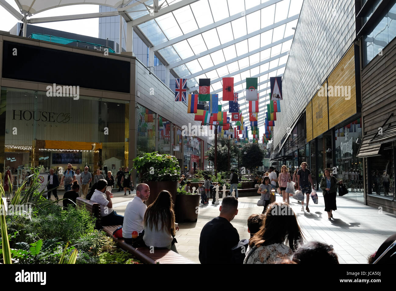 Shopper tragen Shopping Taschen; Menschen auf den Bänken im Einkaufszentrum Westfield Stratford City entspannen; Flaggen der Welt hängen entlang des Korridors Stockfoto