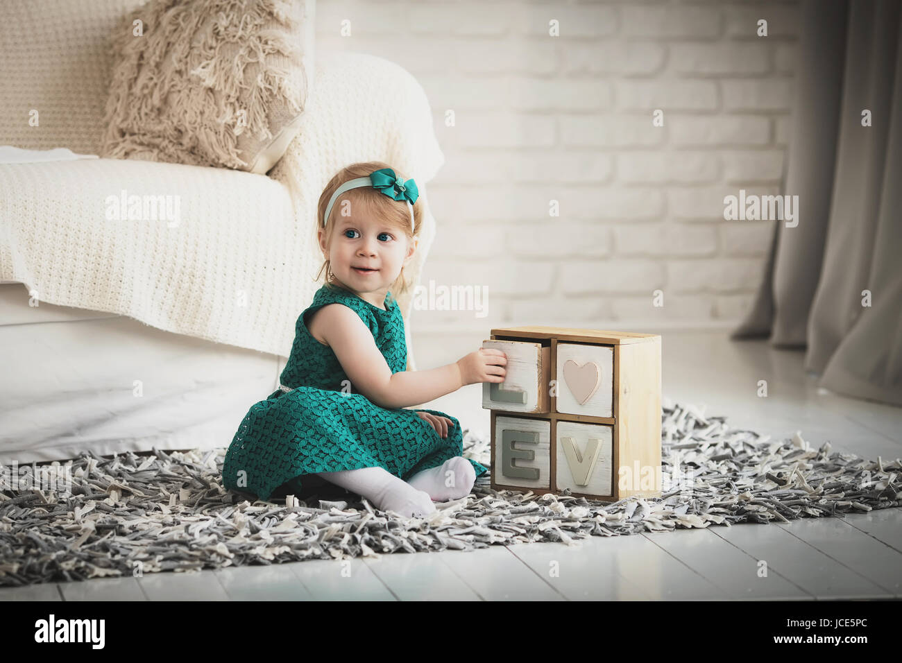 Kind-Modell mit Holzbuchstaben auf grauen flauschiger Teppich drinnen spielen. Kleines Mädchen im grünen Kleid in modernen Wohnung. Stockfoto