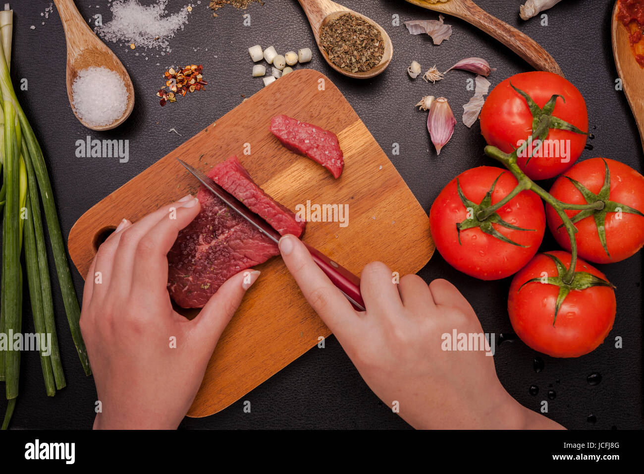 Ein Overhead Foto einer Person, die ein Stück rohes Steak mit Gemüse und anderen Zutaten schneiden. Stockfoto