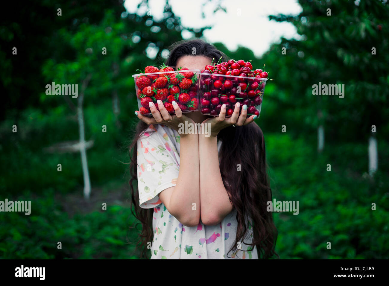 Das Mädchen hält zwei Kisten gefüllt mit Erdbeeren und Kirschen vor dem Hintergrund des Gartens. Konzeption: Wahl Essen vegan Stockfoto