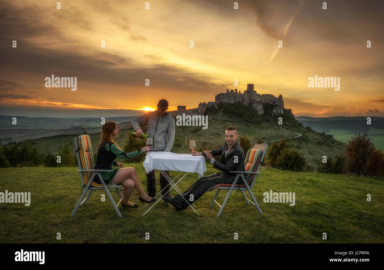 Gruppe von drei Personen trinken Rotwein in der Natur unter den Ruinen einer Burg bei Sonnenuntergang. Stockfoto