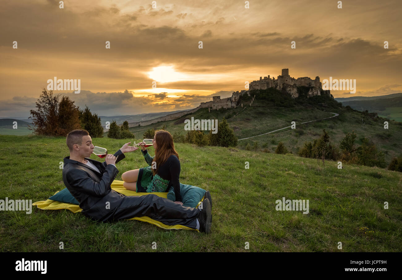 Verliebte Paar auf einer Decke in der Natur liegen und trinken Rotwein unter den Ruinen einer Burg bei Sonnenuntergang. Stockfoto