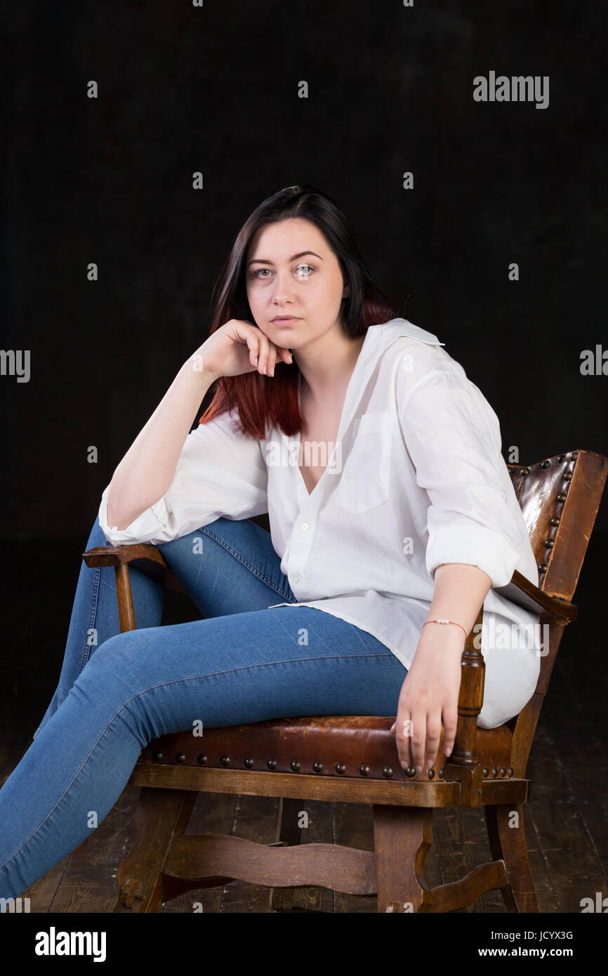 Schöne weiße braune kurzhaarige Hündin mit farbigen in roten enden tragen Männer weißes Hemd und blaue Jeans, sitzt auf einem Vintage Stuhl auf einem dunklen Hintergrund Stockfoto