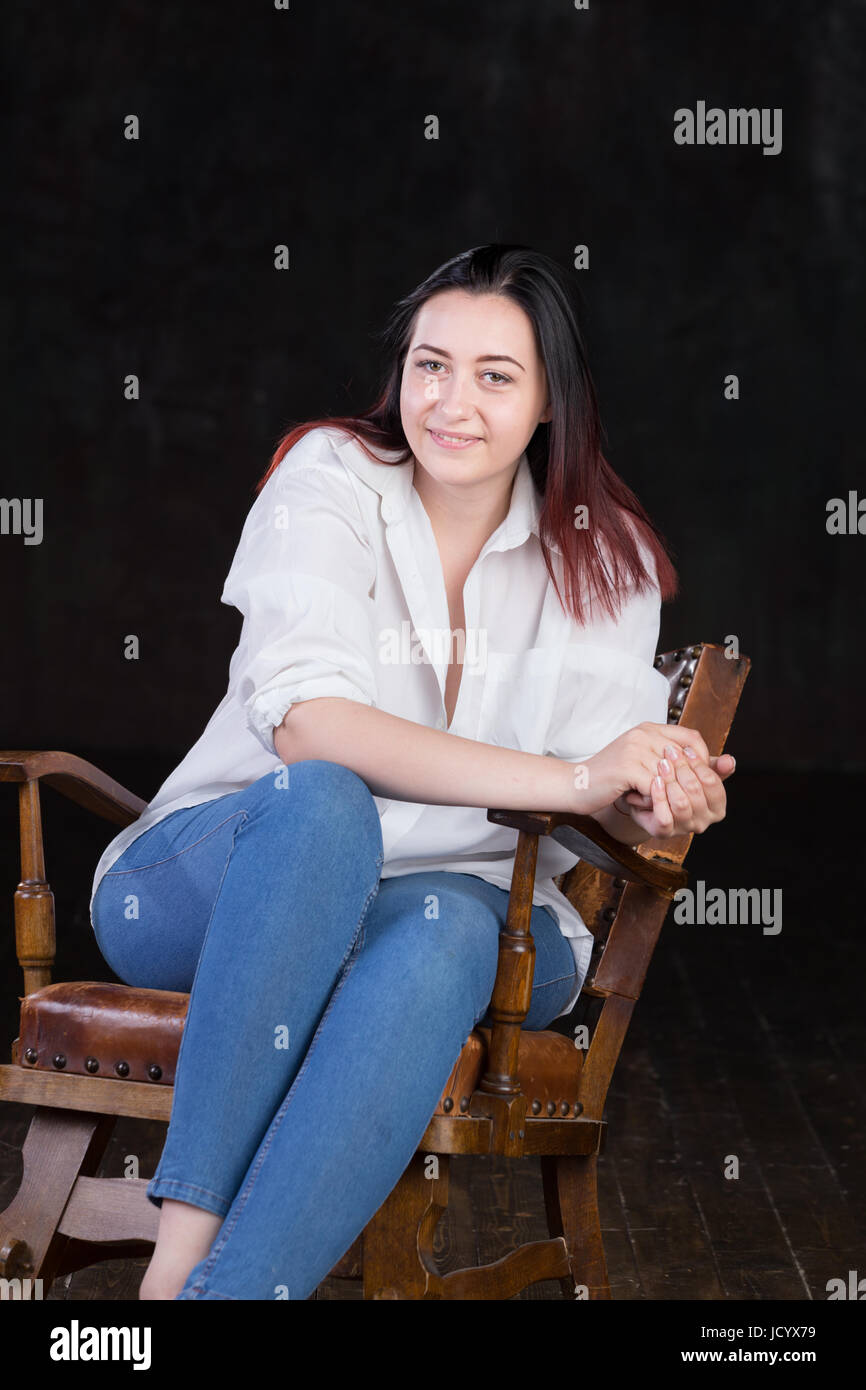 Schöne weiße braune kurzhaarige Hündin mit farbigen in roten enden tragen Männer weißes Hemd und blaue Jeans, sitzt auf einem Vintage Stuhl auf einem dunklen Hintergrund Stockfoto