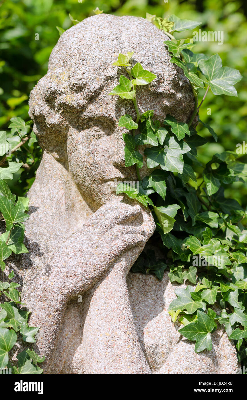 Stein-Statue einer Frau in der Sonne, von Efeu überwuchert. Hedera, Klettern über dem rechten Auge einer alten verwitterten Skulptur mit gefalteten Händen. Stockfoto