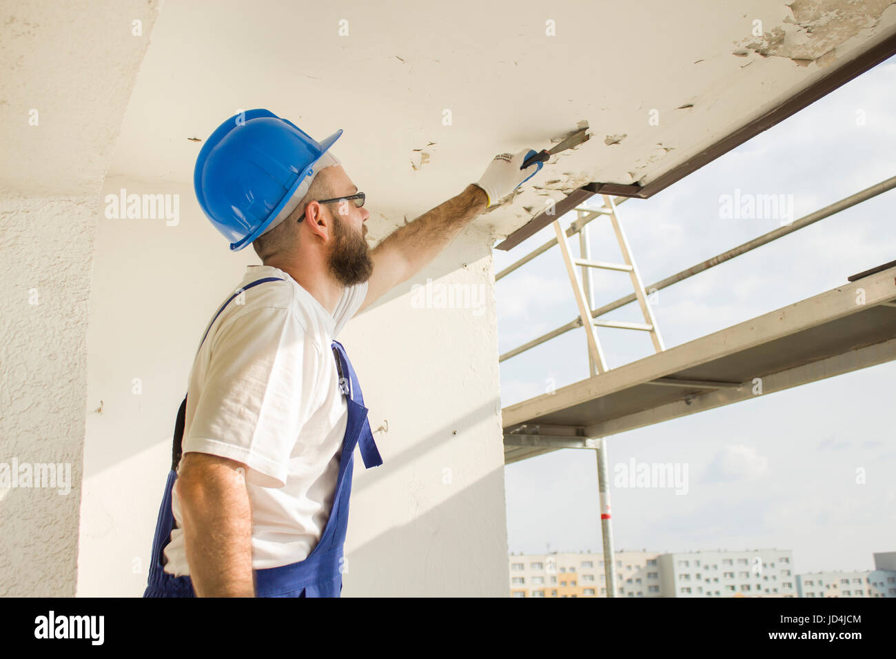 Bauarbeiter in Arbeit Kleidung, Schutzhandschuhe und einen Helm auf. Die alte Farbe Spachtel von der Decke entfernen. Stockfoto