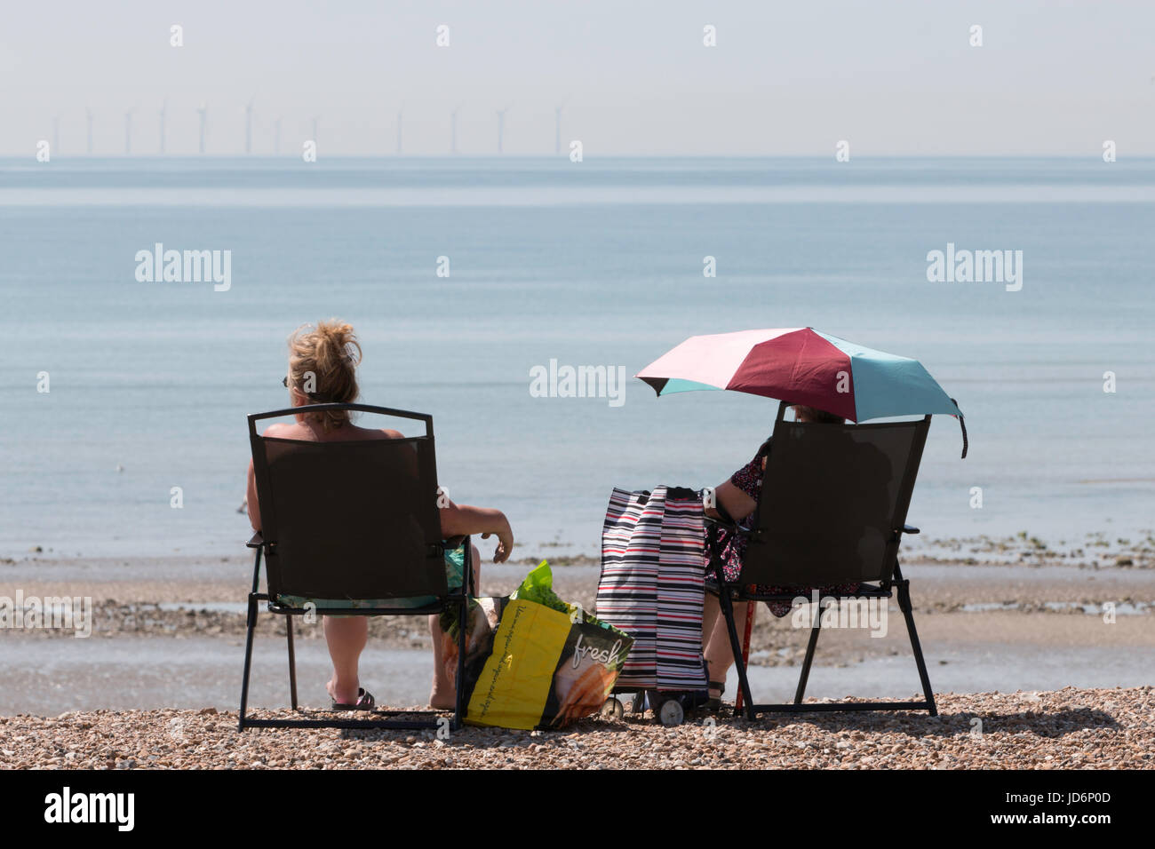 Zwei Damen, die den sommer genießen Sonne am Meer. Man unter einem Sonnenschirm sitzen. Eine Offshore Wind Farm kann am Horizont gesehen werden. Stockfoto