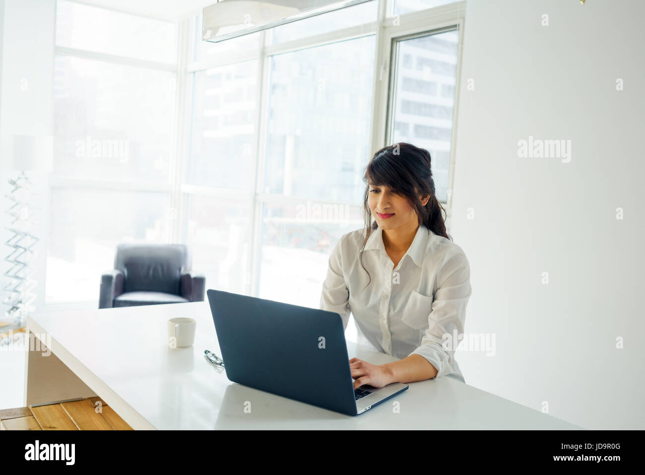 Junge Frau mit Laptop am Schreibtisch, Tag, mit Fenster hinter. junge Erwachsene arabischen hübsche 20 Jahre alt Stockfoto
