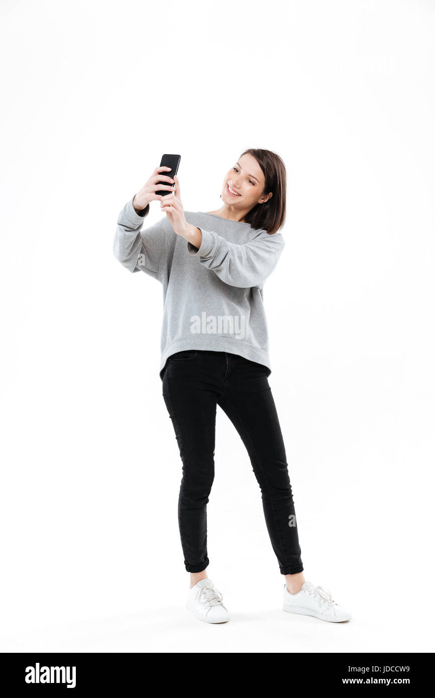 In voller Länge Portrait von einem lächelnden hübschen Mädchen stehen und nehmen Selfie auf Handy isoliert auf weißem Hintergrund Stockfoto