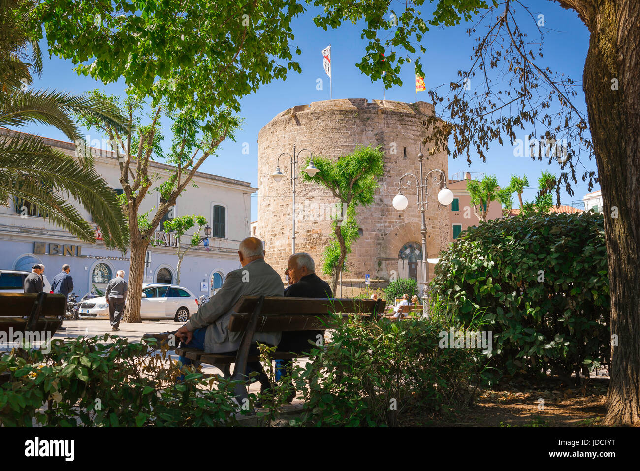 Alte Freunde, Rückansicht von zwei älteren Männern, die auf einer Bank in öffentlichen Gärten in der Nähe des mittelalterlichen Torre di Porta Terra in der Altstadt von Alghero, Sardinien, plaudern Stockfoto