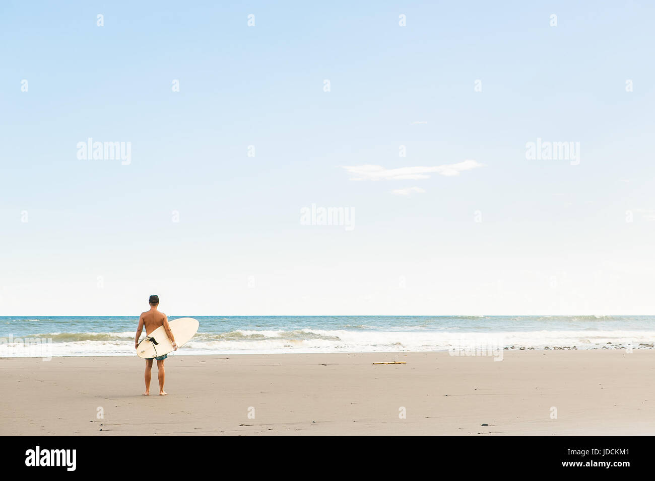 Gut aussehender Mann in blau Badeshorts und Mütze mit langen Surf Board warten auf Surfspot am Meer Ozeanstrand Surfen stehen. Weiße leere Surfbrett. Konzept-o Stockfoto