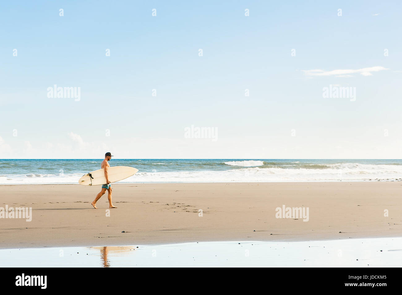 Gut aussehender Mann in blau Badeshorts und Kappe gehen mit lange Surf Surf Board warten auf Surfspot am Meer Ozeanstrand. Weiße leere Surfbrett. Konzept der Stockfoto