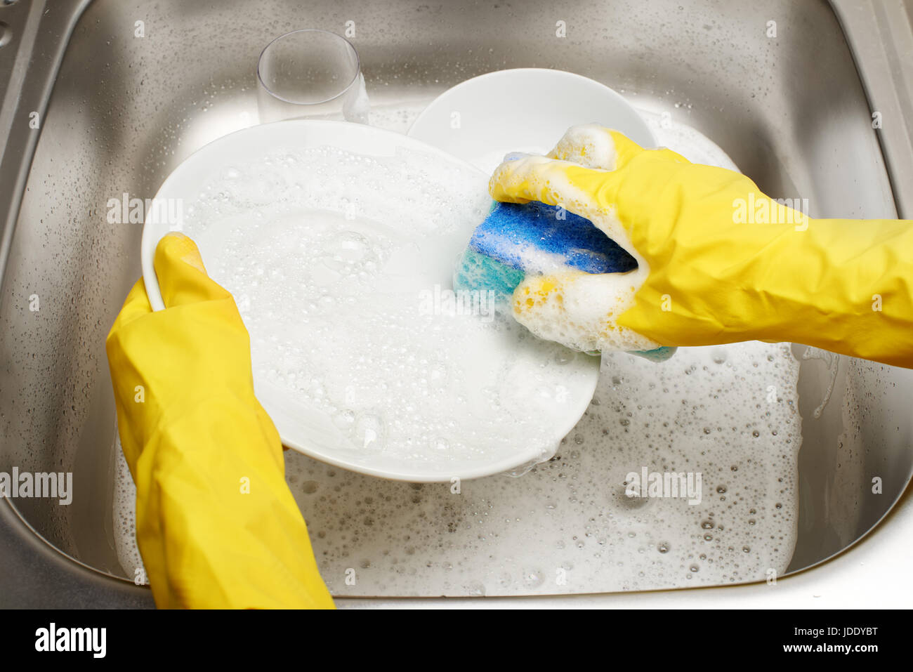 Nahaufnahme von weiblichen Händen gelb Gummischutz Handschuhe waschen weißen Teller mit blauen Reinigung gegen Küchenspüle voller Schaum und Tablew Schwamm Stockfoto