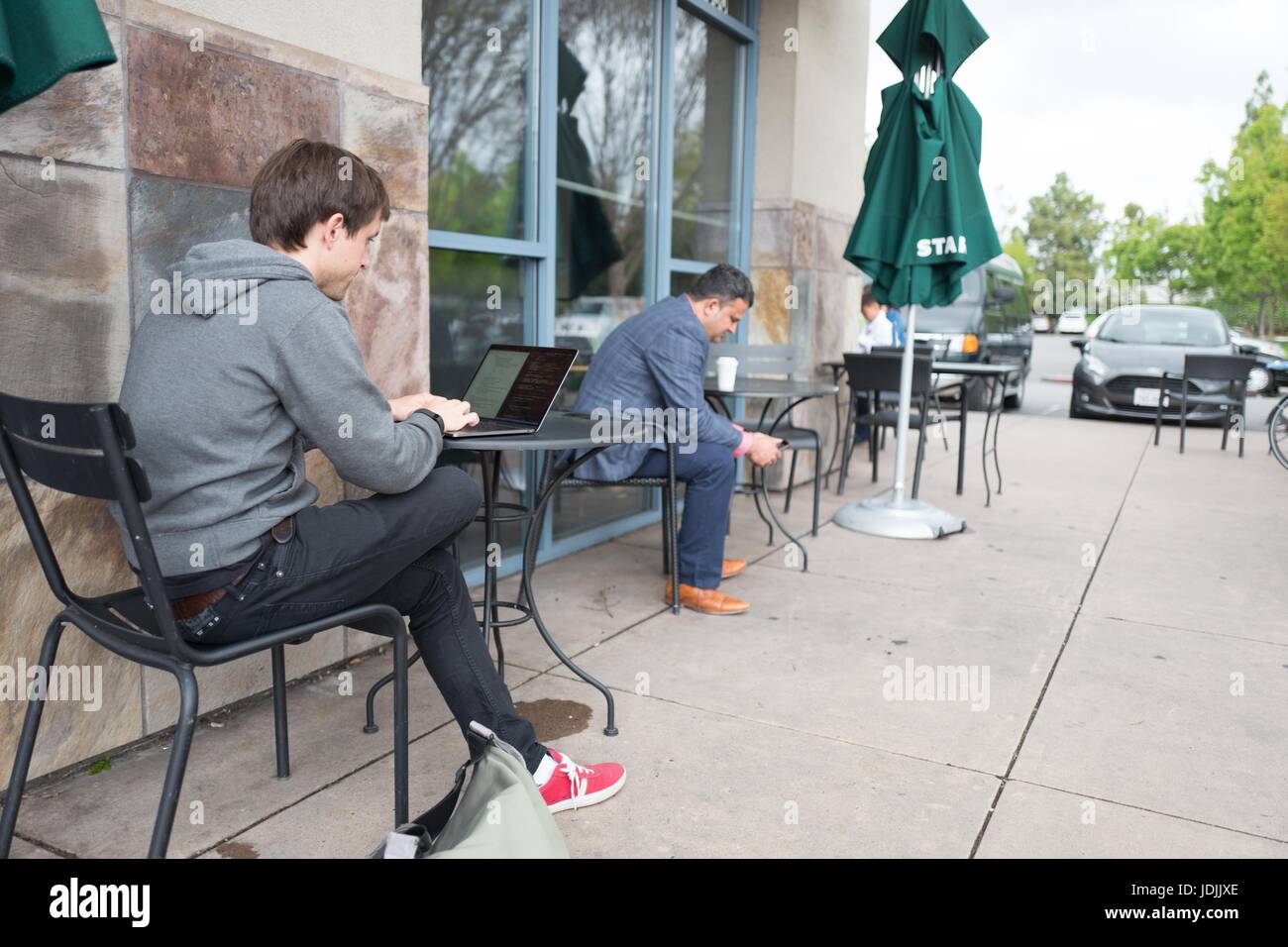 Ein Junge, tausendjährigen Alter männlicher Tech Arbeiter in einem Kapuzenpullover im Freien sitzt und arbeitet auf einem Laptop in der Nähe der Googleplex Hauptquartier von Google Inc in das Silicon Valley Stadt Mountain View, Kalifornien, 7. April 2017. Stockfoto