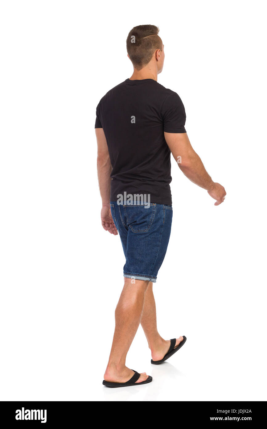 Junger Mann zu Fuß in schwarzes t-Shirt, Jeans-Shorts und schwarze Flip-flops. Ansicht der Rückseite. Voller Länge Studio gedreht isoliert auf weiss. Stockfoto