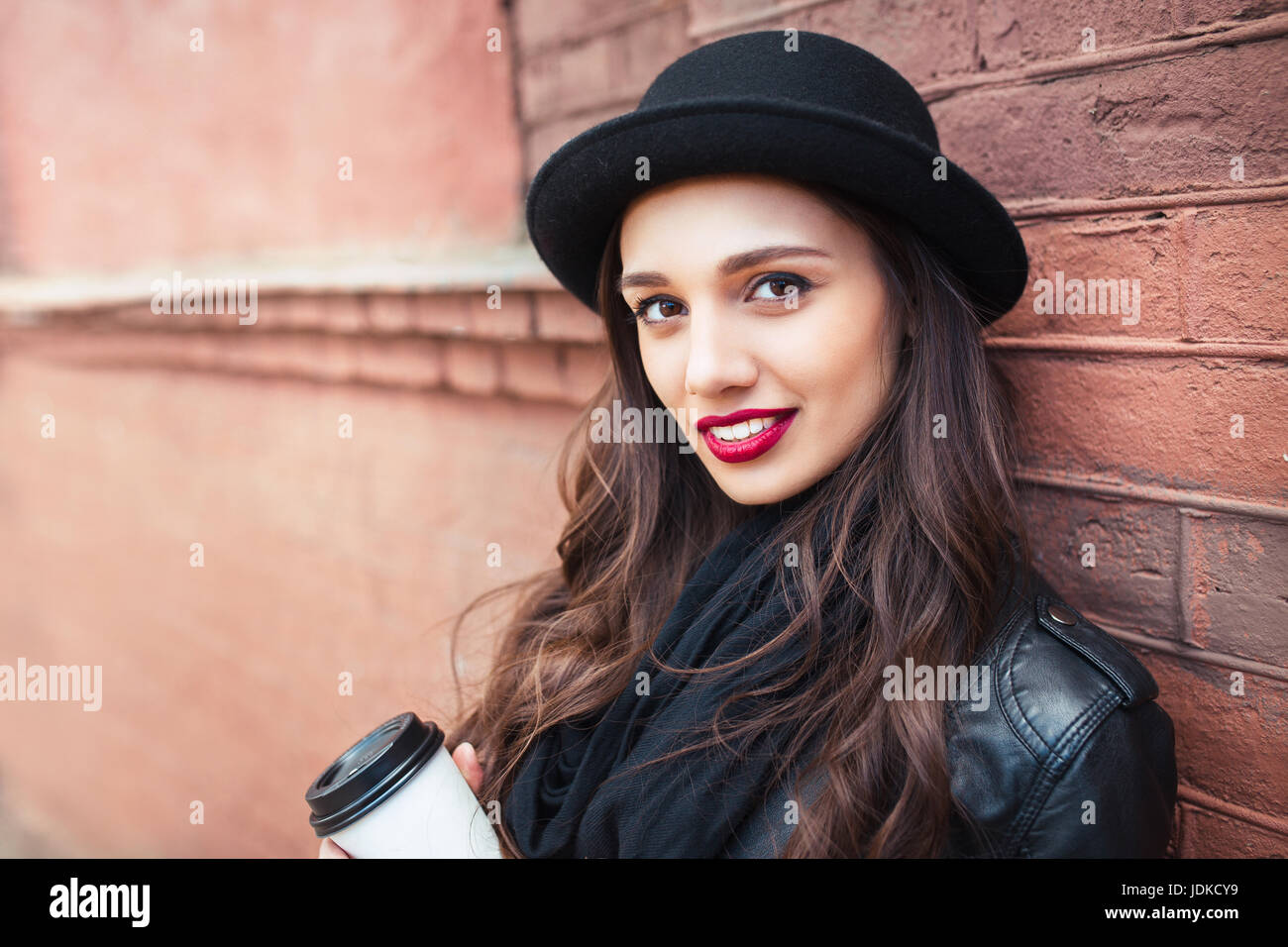 junge hübsche Brünette, trinken Kaffee herumlaufen city.leather Jacke, städtische Rucksack, knallrote Lippen und Hut Stockfoto