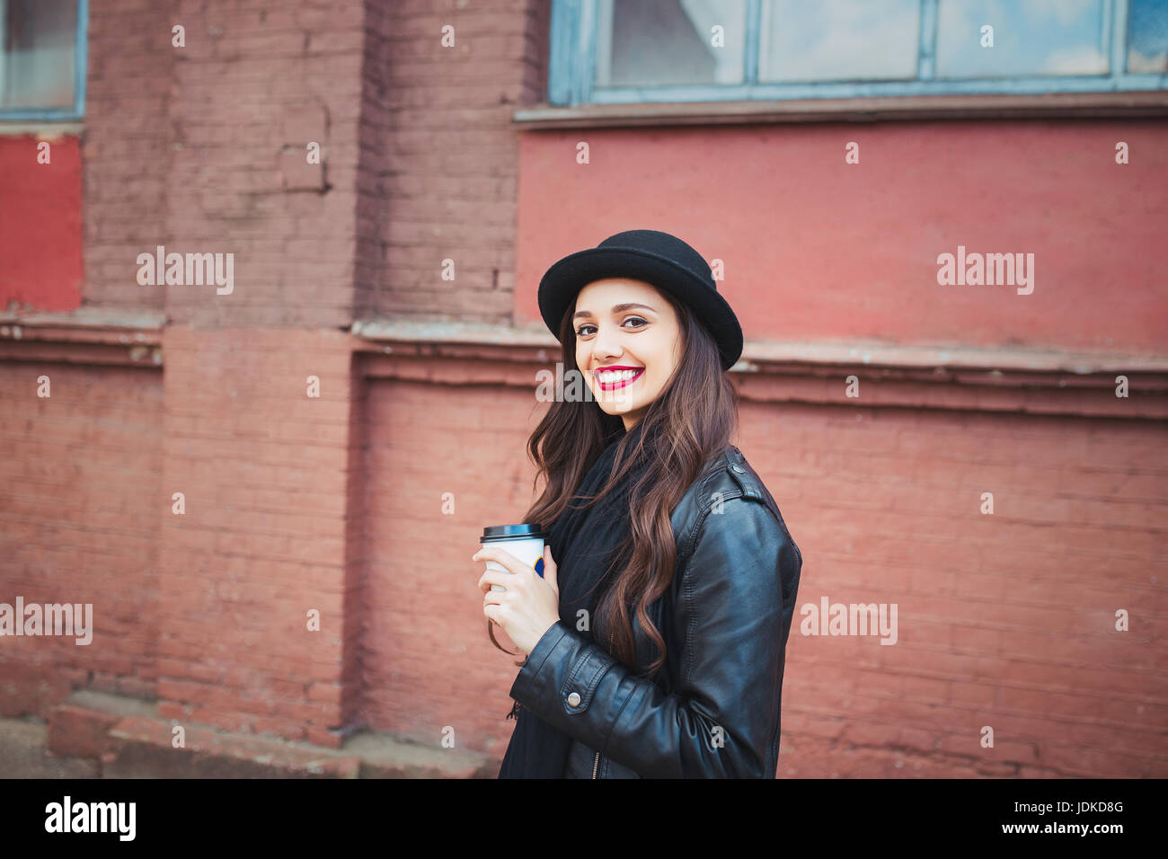 junge hübsche Brünette, trinken Kaffee herumlaufen city.leather Jacke, städtische Rucksack, knallrote Lippen und Hut Stockfoto