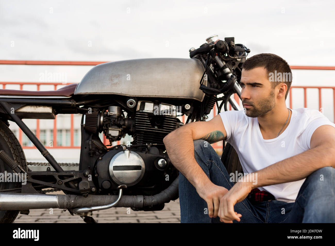 Gut aussehend glücklicher Fahrer Mann mit Bart und Schnurrbart in der Nähe von seinem klassischen Stil Biker Café Racer Motorrad sitzen und schauen Sie auf der Seite. Fahrrad benutzerdefinierte gemacht in vint Stockfoto