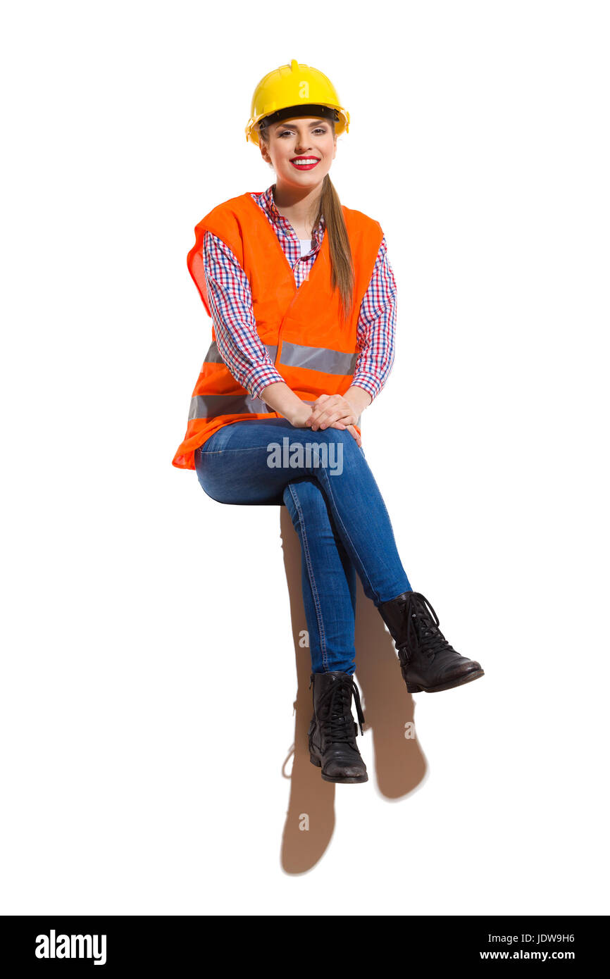 Lächelnde junge Frau in gelben Bauarbeiterhelm, orange Warnweste, Holzfällerhemd, Jeans und schwarzen Stiefeln, sitzend mit Beine gekreuzt, oben und auf der Suche ein Stockfoto
