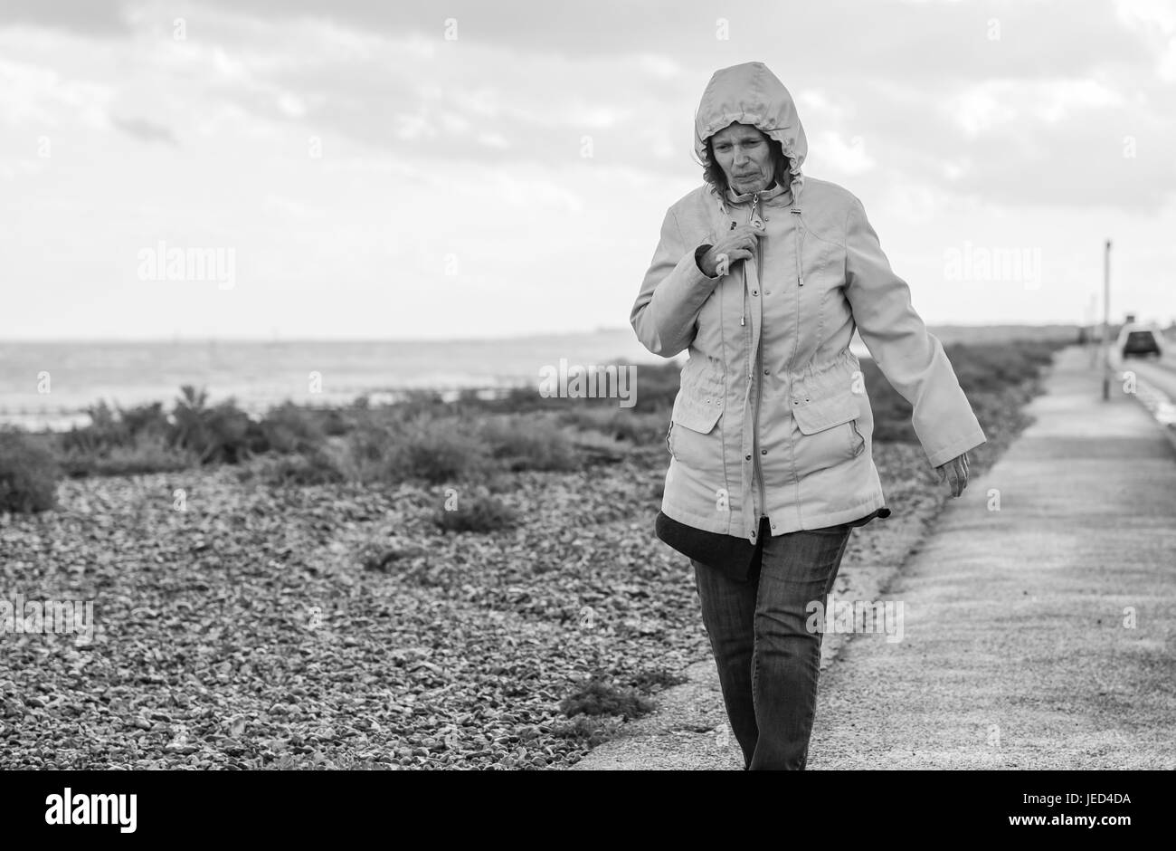 Nass und windig. Dame zu Fuß entlang einer Küstenstraße in den Wind an einem trüben Tag. Schwarz / weiß Bild. Stockfoto