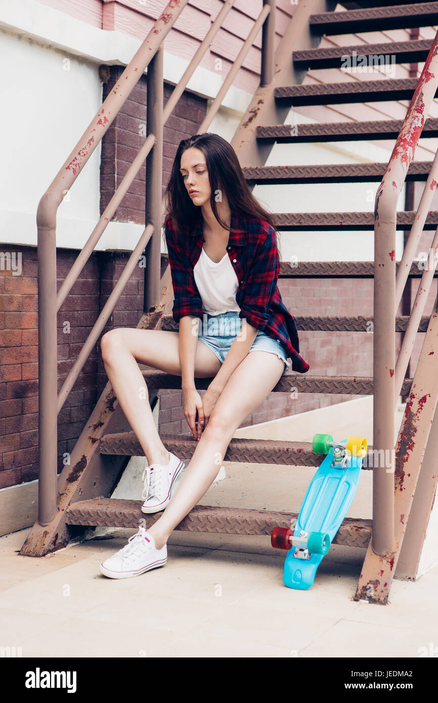 Junge schöne Mädchen in karierte Hipster Shirt und kurze Jeans Shorts und  weiße Turnschuhe sitzen auf der Treppe mit ihren blauen Mini Skateboard  Longboard. U Stockfotografie - Alamy