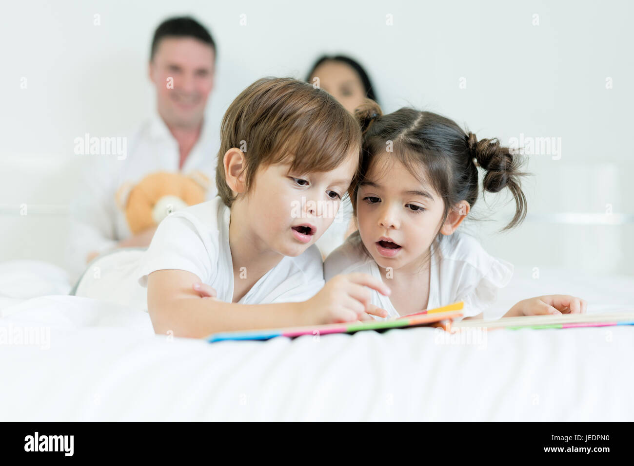 Asiatischen Brüder und asiatische Tochter Lesebuch zusammen auf dem Bett im Schlafzimmer mit Vater und Mutter im Hintergrund. Glückliche Familien-Konzept. Stockfoto