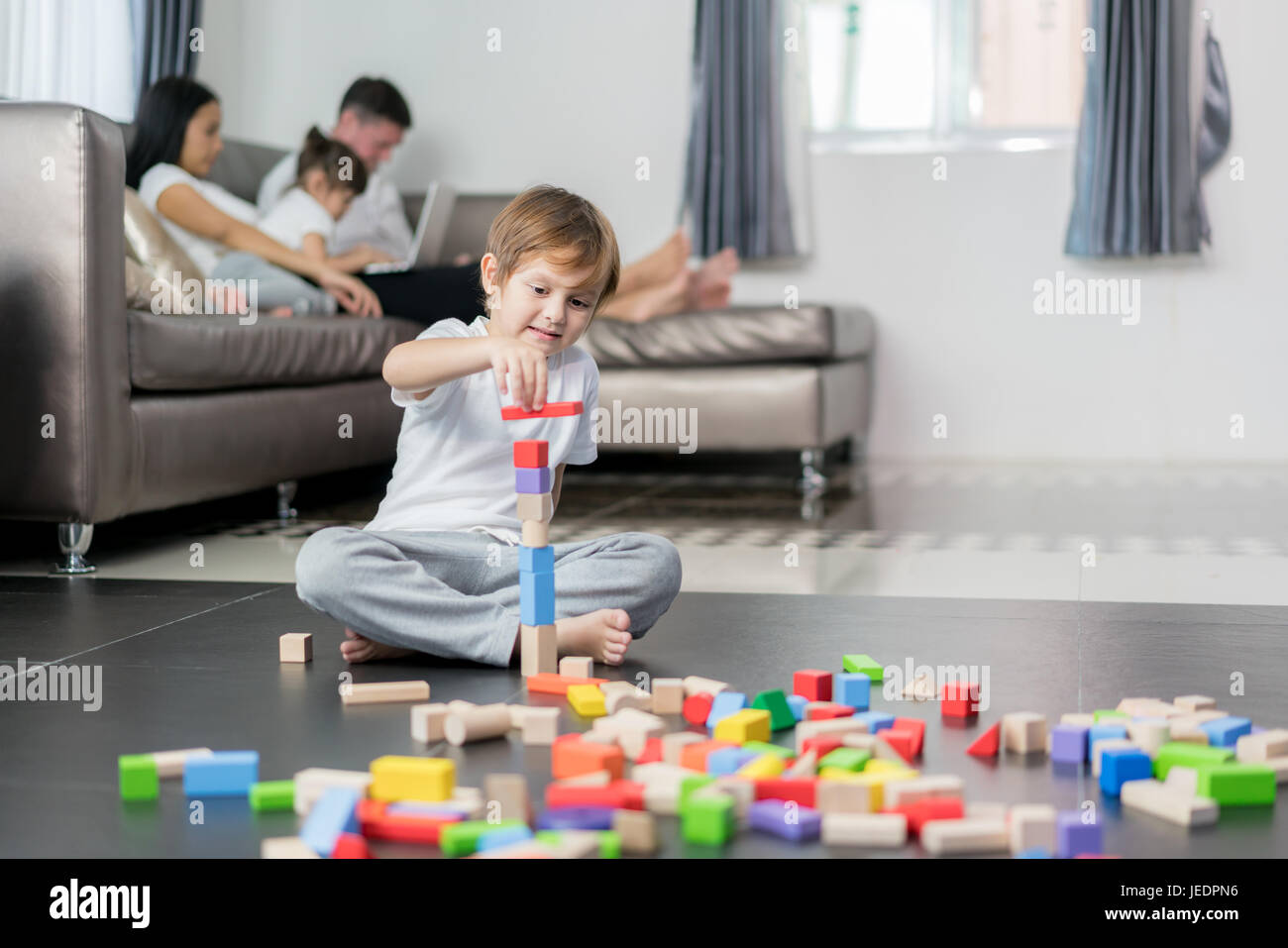 Asiatische junge Holz Spielzeug im Wohnzimmer mit Vater, Mutter und Tochter im Hintergrund zu spielen. Glückliche Familie. Stockfoto