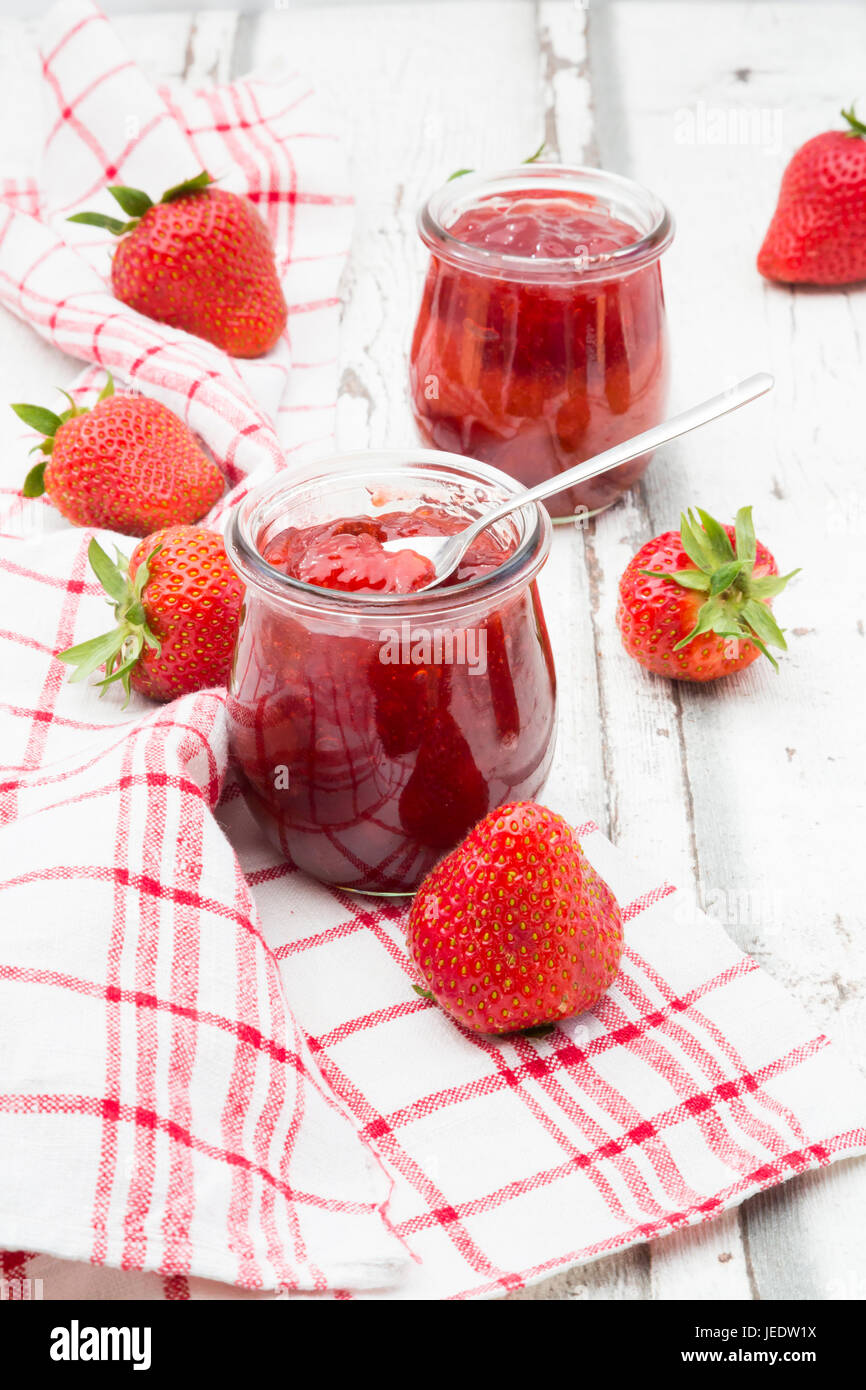 Zwei Gläser hausgemachte Erdbeermarmelade, Küchentuch und Erdbeeren auf weißem Holz Stockfoto