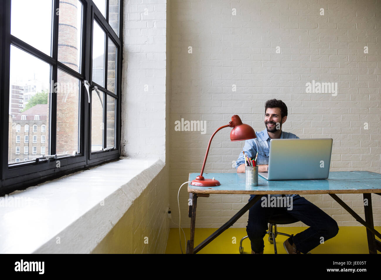 Lächelnder junge Mann sitzt am Schreibtisch mit Laptop Blick durch Fenster Stockfoto