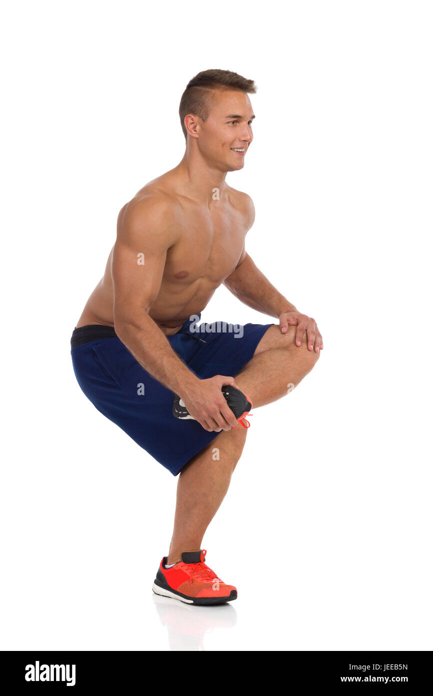 Junge fit Mann in blauen Hosen und orange Sneakers hocken auf einem Bein, hält sein Bein auf andere Bein Knie und stretching Gluteus Maximus. Seitenansicht. Fu Stockfoto