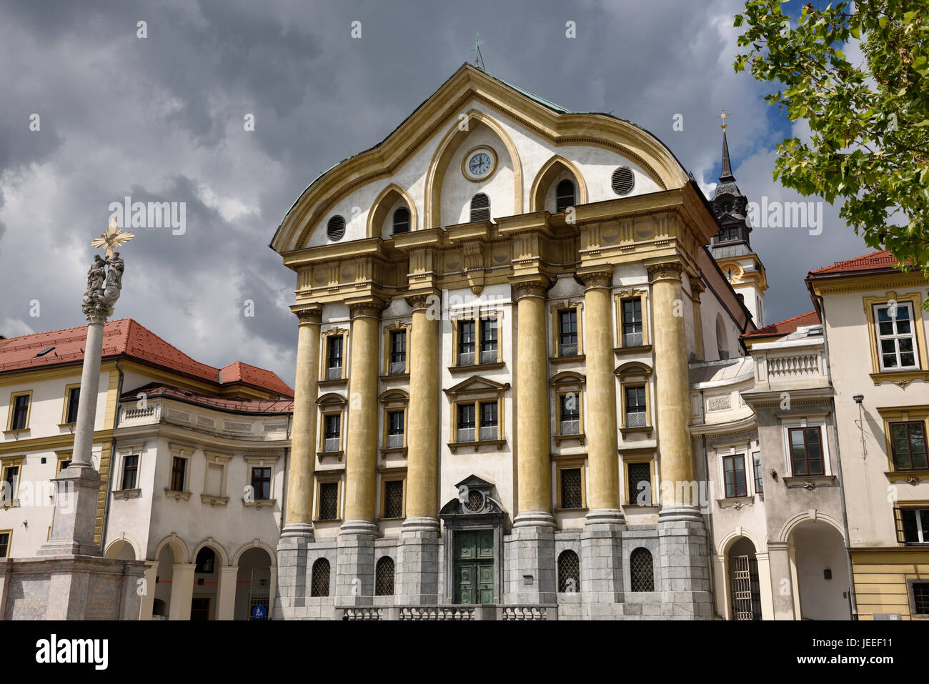 Volle Sonne auf die Ursulinen-Kirche der Heiligen Dreifaltigkeit mit Marmor-Statuen der Heiligen Dreifaltigkeit Spalte in Ljubljana Slowenien mit dunklen Wolken Stockfoto