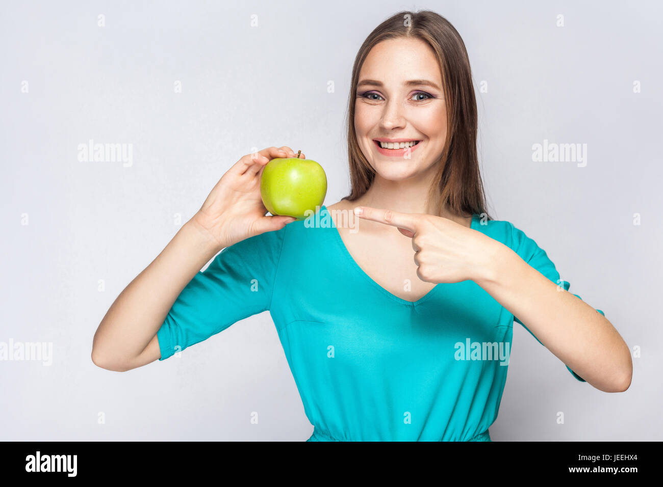 Junge schöne Frau mit Sommersprossen und grünes Kleid hält Apfel und deutete mit dem Finger. Studioaufnahme, isoliert auf hellgrauen Hintergrund. Stockfoto