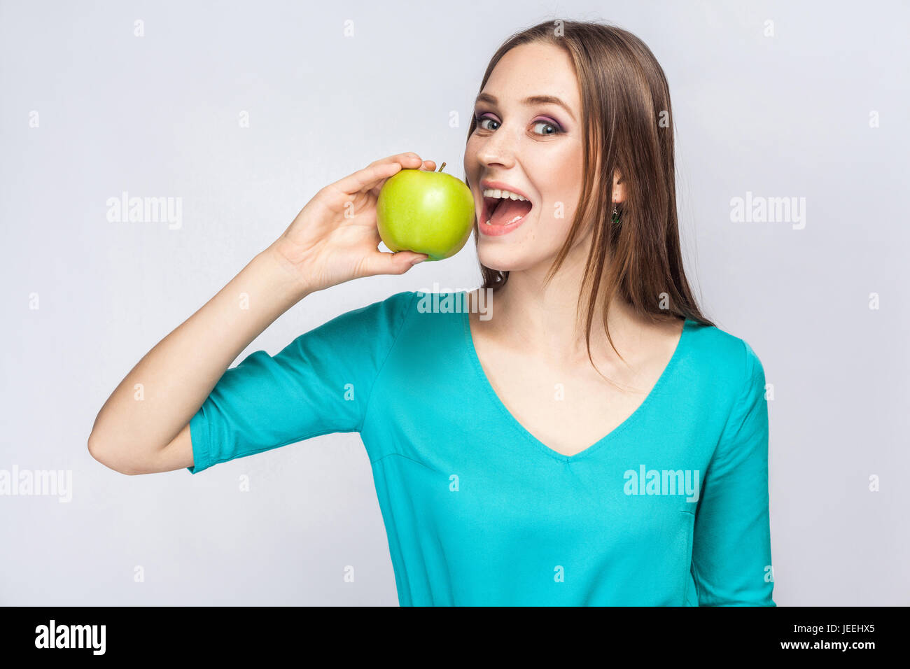Junge schöne Frau mit Sommersprossen und grünes Kleid hält Apfel und Essen, Blick in die Kamera. Studioaufnahme, isoliert auf hellgrauen Hintergrund. Stockfoto