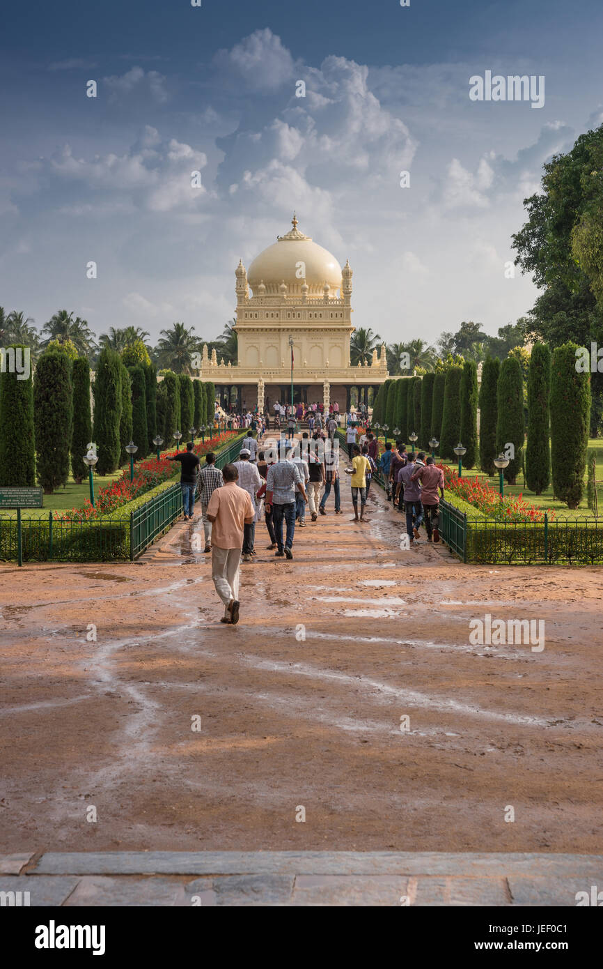 Mysore, Indien - 26. Oktober 2013: Grüne und rote Park mit Weg zu Creme gelb Tipu Sultan Mausoleum mit Kuppel unter schweren Wolkengebilde. Menschen walki Stockfoto