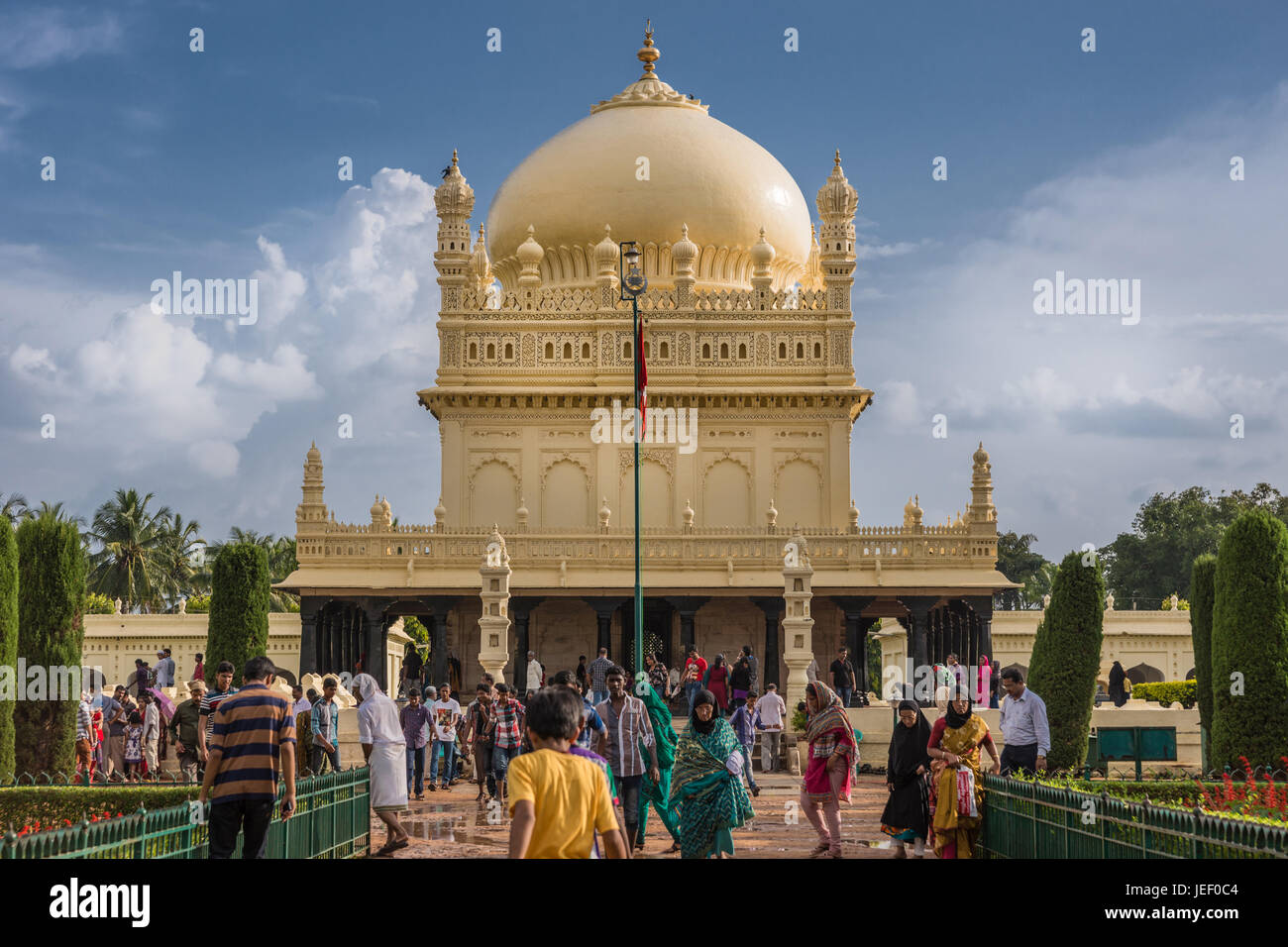 Mysore, Indien - 26. Oktober 2013: Creme gelb Tipu Sultan Mausoleum mit Kuppel unter schweren Wolkengebilde. Menschen zu Fuß, und aus dem Gebäude. Grün Stockfoto