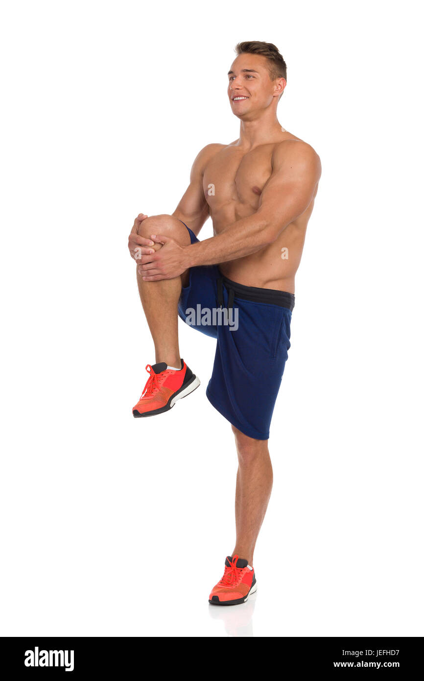 Junge fit Mann in blauen Hosen und orange Sneakers stehen auf einem Bein, hält seine Knie und stretching Gluteus Maximus. Seitenansicht. Voller Länge Studio s Stockfoto