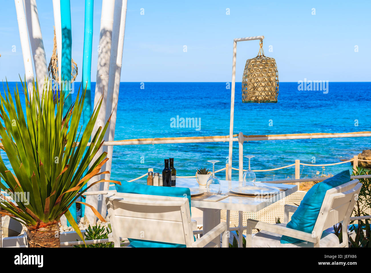 Weißen Tisch und Stühlen mit blauen Kissen auf Restaurant-Terrasse am Strand von Cala Nova mit wunderschönem Blick aufs Meer, Insel Ibiza, Spanien Stockfoto