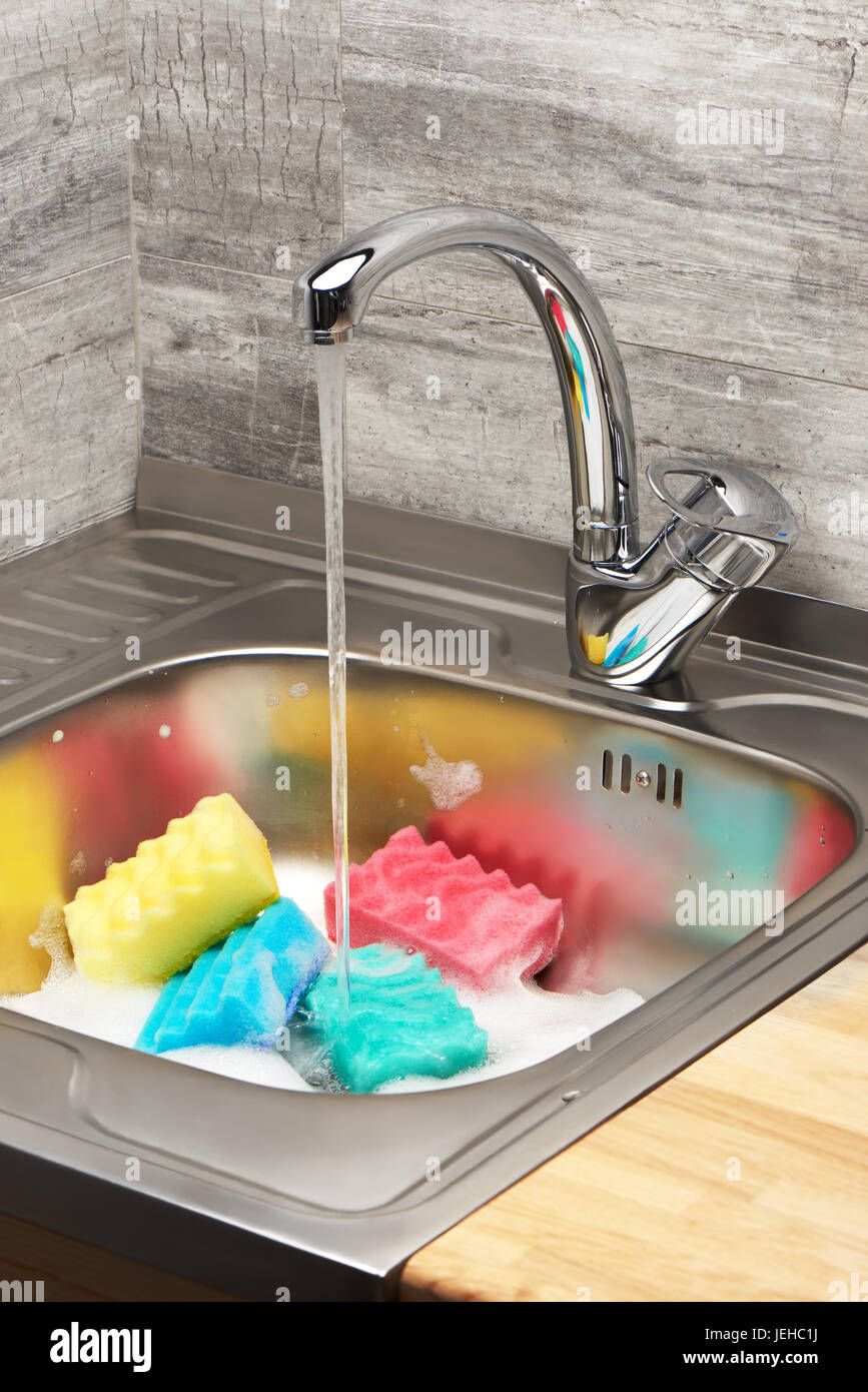 Nahaufnahme der Küchenspüle voller Schaum mit fließendem Leitungswasser, blau, Pink, gelb und grün Reinigung Schwämme Stockfoto