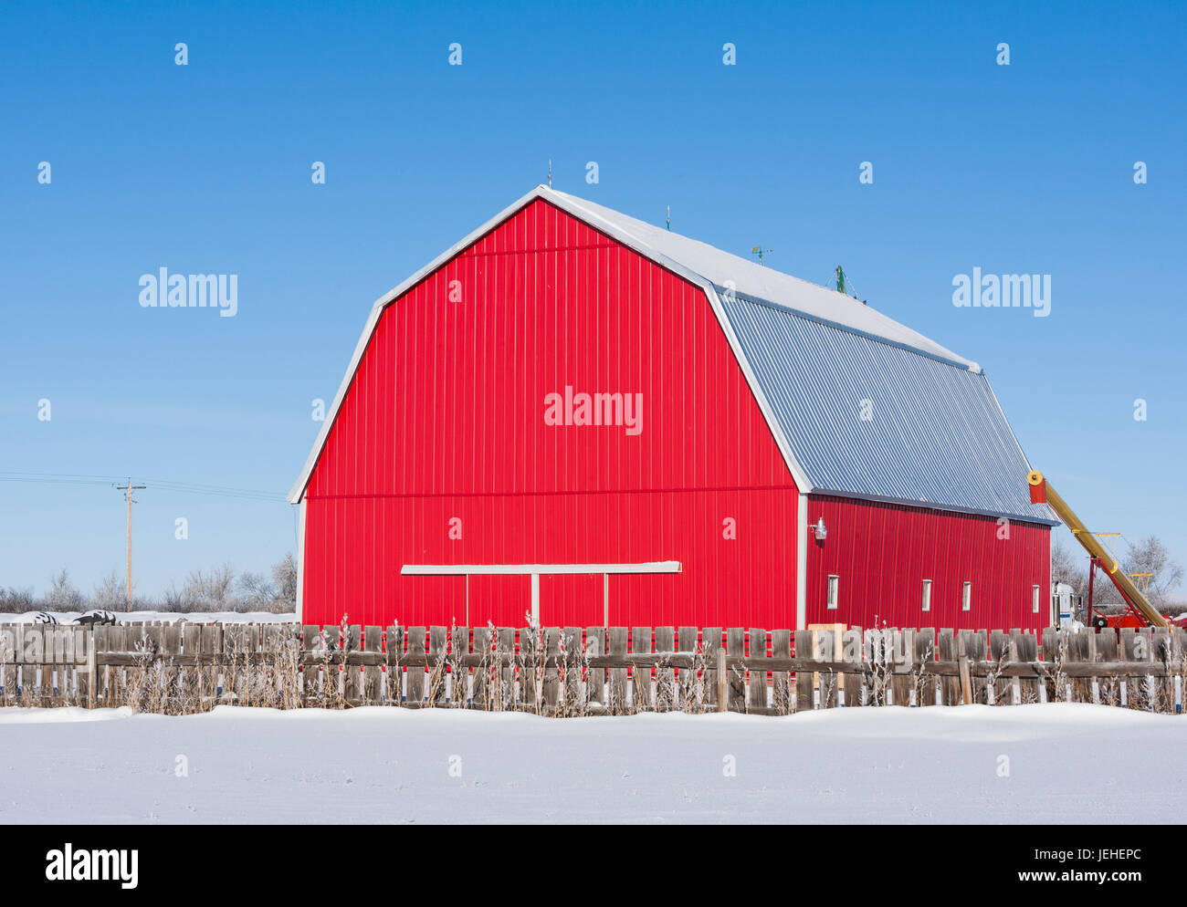 Eine helle rote Scheune ein schneebedecktes Feld mit Holzzaun im Vordergrund, unter blauem Himmel; Alberta, Kanada Stockfoto