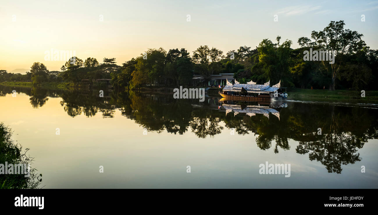 Reflexionen in einem ruhigen Fluss bei Sonnenuntergang mit einem Ausflugsboot entlang der Küste; Chiang Rai, Thailand Stockfoto