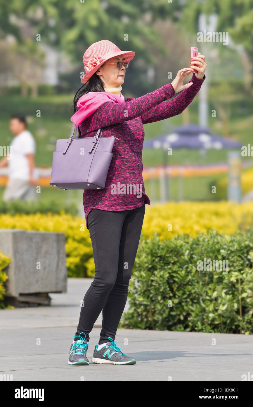 PEKING, 28. APRIL 2016. Die modische Frau mittleren Alters nimmt an einem sonnigen Tag Selfie mit ihrem Smartphone in einem Park auf. Stockfoto