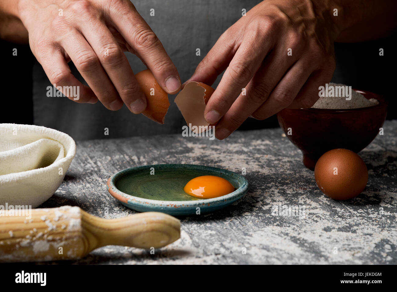 Closeup von einem kaukasischen Jüngling trägt eine graue Schürze krachend ein Ei auf einem rustikalen Holztisch mit Mehl Neben einem Nudelholz bestreut Stockfoto