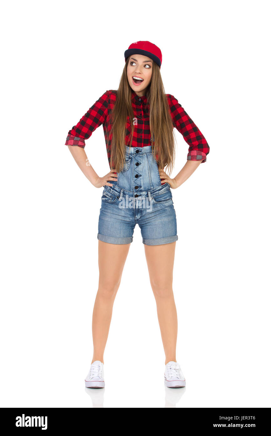 Frau in Rot lumberjack Shirt, Jeans latzhosen Shorts, weiße Turnschuhe und fullcap stand mit offenem Mund, Hände auf Hüfte und blickte. Volle l Stockfoto