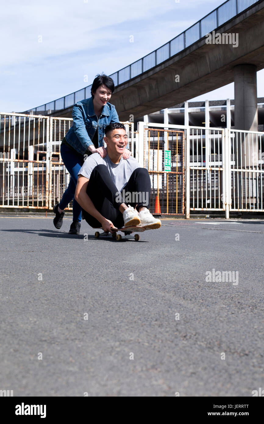 Junge Paare, die Spaß in der Stadt. Die Frau treibt ihr Partner entlang, als er auf einem Skateboard sitzt. Sie lachen beide. Stockfoto