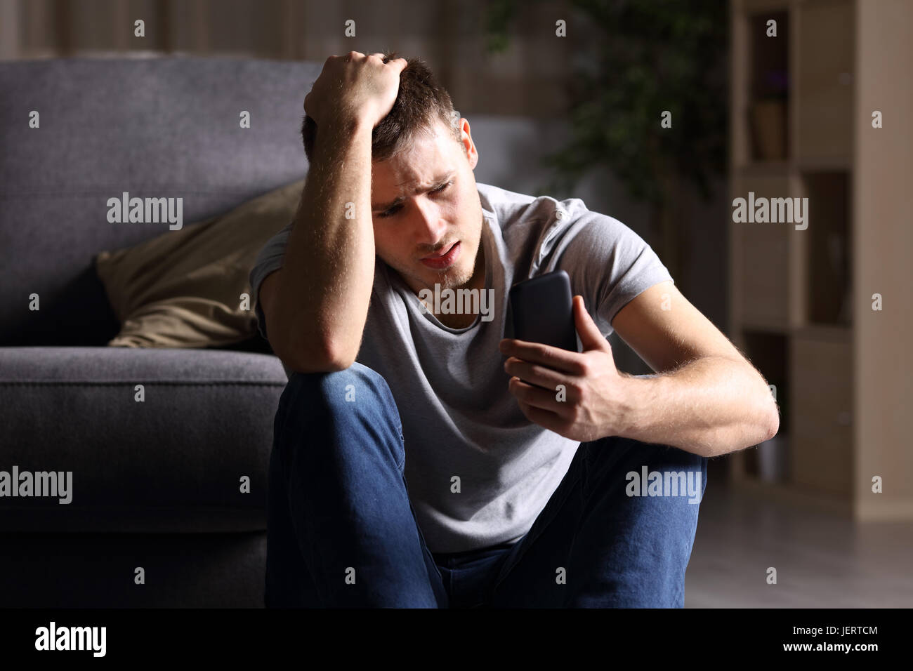 Einzelne trauriger Mann Handy sitzen auf dem Boden im Wohnzimmer zu Hause mit einem dunklen Hintergrund prüfen Stockfoto