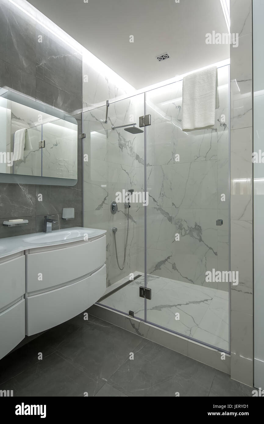 Hellen Badezimmer mit strukturierten gefliesten Wänden. Es gibt eine Dusche mit einem Glas Tür, weißen Waschbecken mit einem Wasserhahn, Spiegel, leuchtenden Lampen, Handtuch aufhängen. Indoor Stockfoto