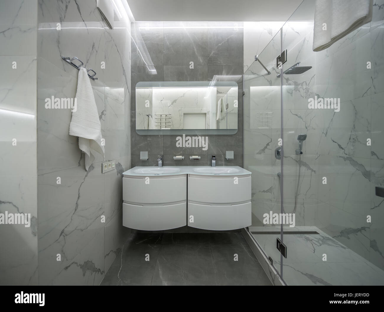 Modernes Bad mit Licht geflieste Wände. Es gibt eine Dusche mit Glastür, zwei weiße Waschbecken mit Armaturen, Spiegel über Ihnen, leuchtenden Lampen, Aufhänger wi Stockfoto